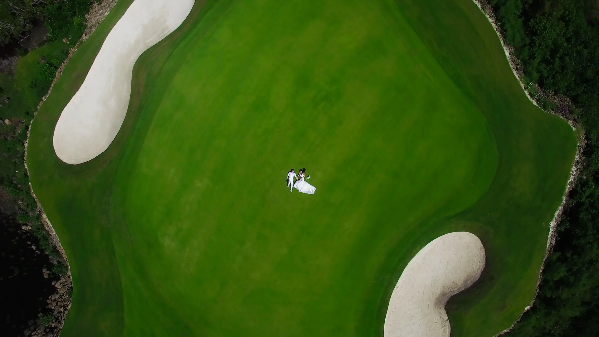 An aerial view of a bride and groom lying on a golf green beside two bunkers